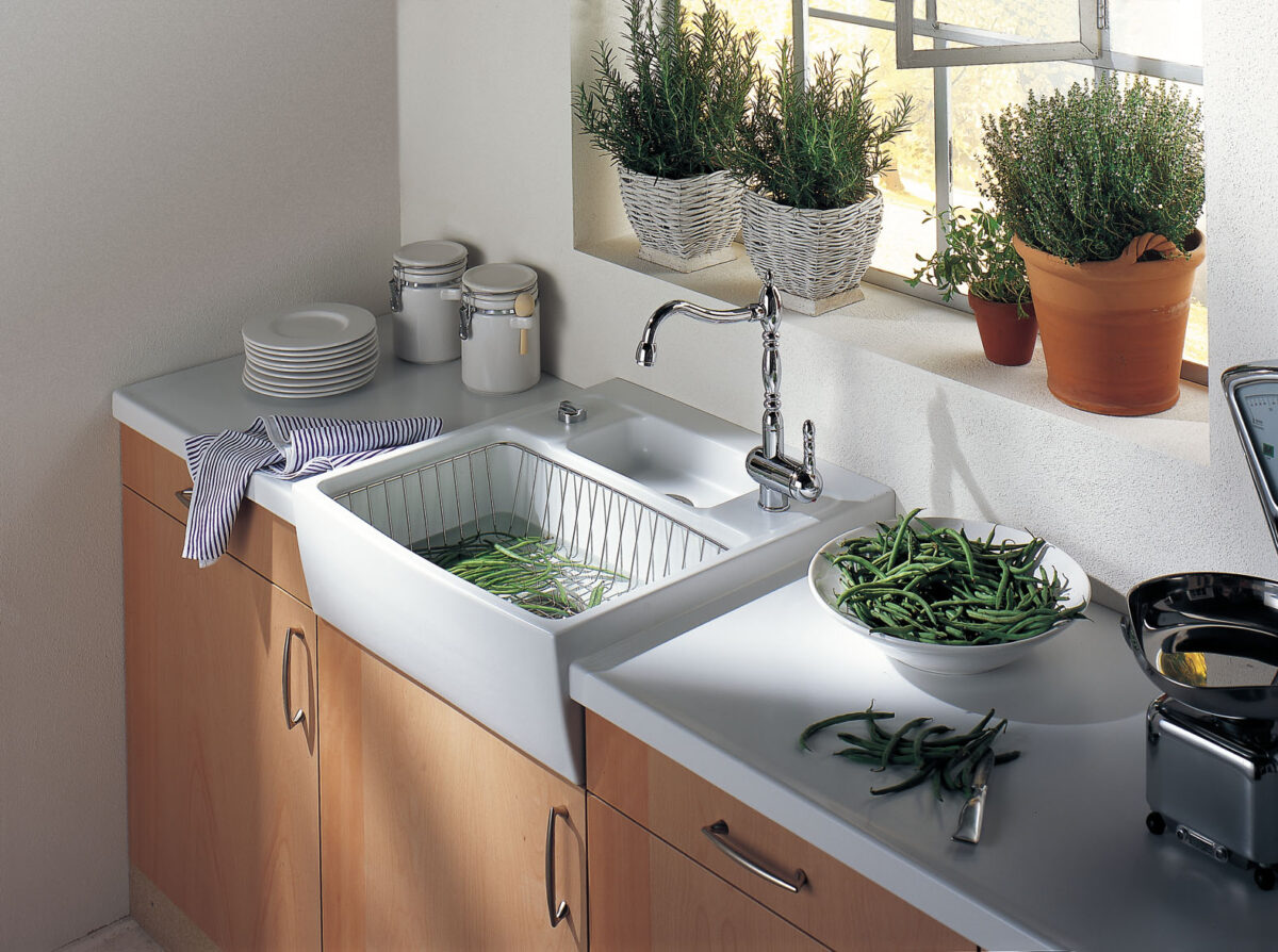 White ceramic farmhouse sink by Systemceram with wooden cabinets and fresh herbs on the windowsill.